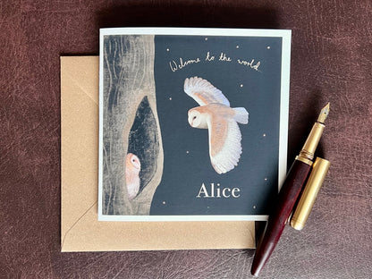Personalised Owl New Baby Greeting Card (Premium Card) laid flat on a recycled kraft envelope, featuring a barn owl flying to its baby in a woodland night scene with handwritten text “Welcome to the World” and the personalised name at the bottom, on a matte black nighttime background, styled on a deep brown leatherette surface with a wooden fountain pen