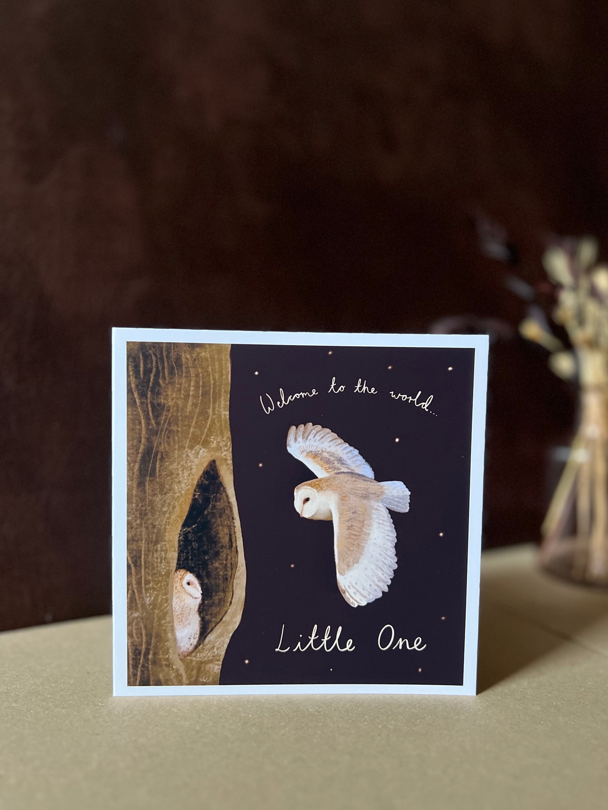Personalised Owl New Baby Greeting Card (Standard Card) standing on a kraft paper surface with a deep brown leatherette background, showing the front illustration of a barn owl flying to its baby in a woodland night scene with handwritten text “Welcome to the World, Little One”; styled with a vase and dried flowers out of focus to create a natural woodland home display
