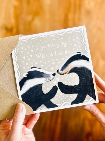 Hands holding personalised woodland wedding card with illustrated badgers, removed from kraft envelope, sage green design with couple names on front