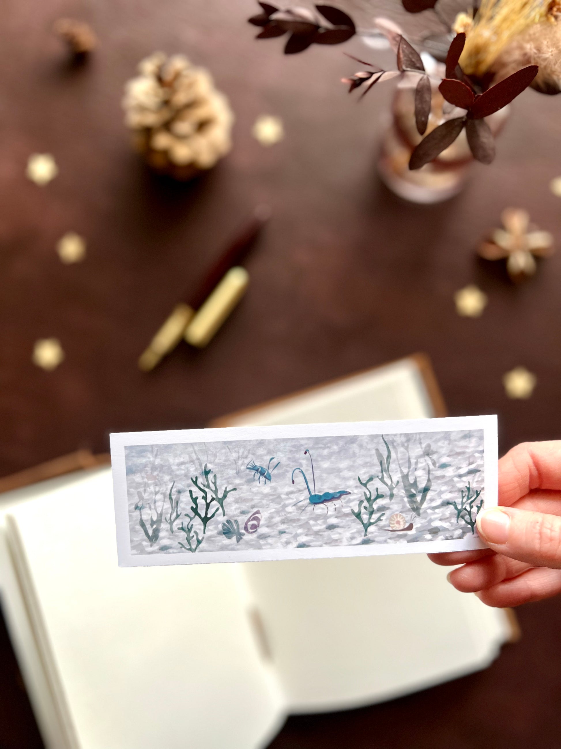 Illustrated bookmark featuring a prawn and lobster on a white pebble seabed in a calm ocean scene. Woodland themed background with sketchbook, pine cones, and kraft paper stars. Part of a 16 pack animal bookmark set for wedding favours.