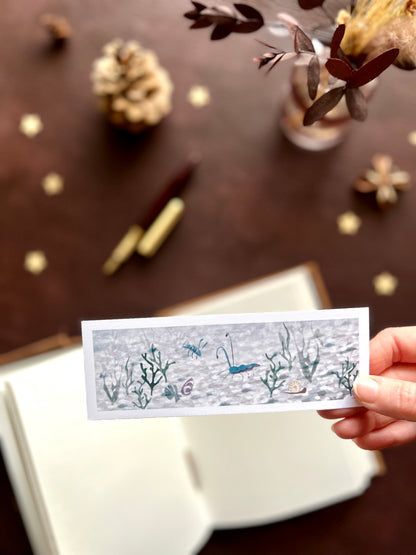 Illustrated bookmark featuring a prawn and lobster on a white pebble seabed in a calm ocean scene. Woodland themed background with sketchbook, pine cones, and kraft paper stars. Part of a 16 pack animal bookmark set for wedding favours.