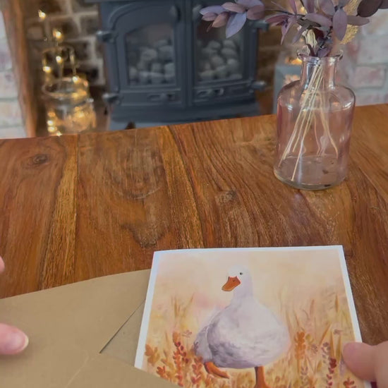 Video showing the Duck in the Meadow birthday card being removed from its brown, recycled kraft envelope and opened to reveal a blank interior. Demonstrates personalised text options on the front, card standing on a rustic wooden table with farmhouse decor, and shows the back with product and artist information. Handcrafted illustration of a white duck in orange flowers for a whimsical, heartwarming greeting card.”