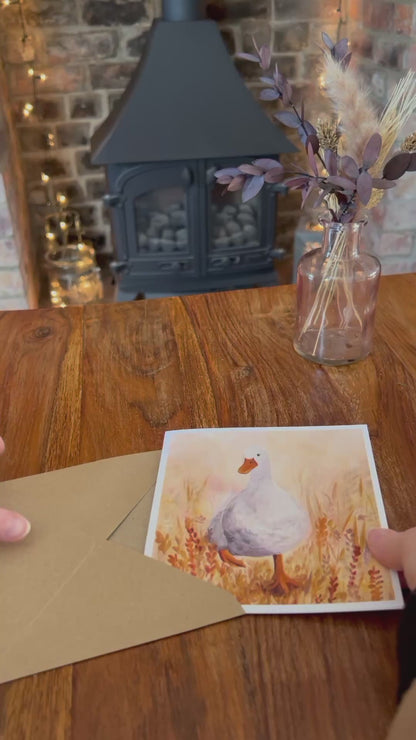 Video showing the Duck in the Meadow birthday card being removed from its brown, recycled kraft envelope and opened to reveal a blank interior. Demonstrates personalised text options on the front, card standing on a rustic wooden table with farmhouse decor, and shows the back with product and artist information. Handcrafted illustration of a white duck in orange flowers for a whimsical, heartwarming greeting card.”