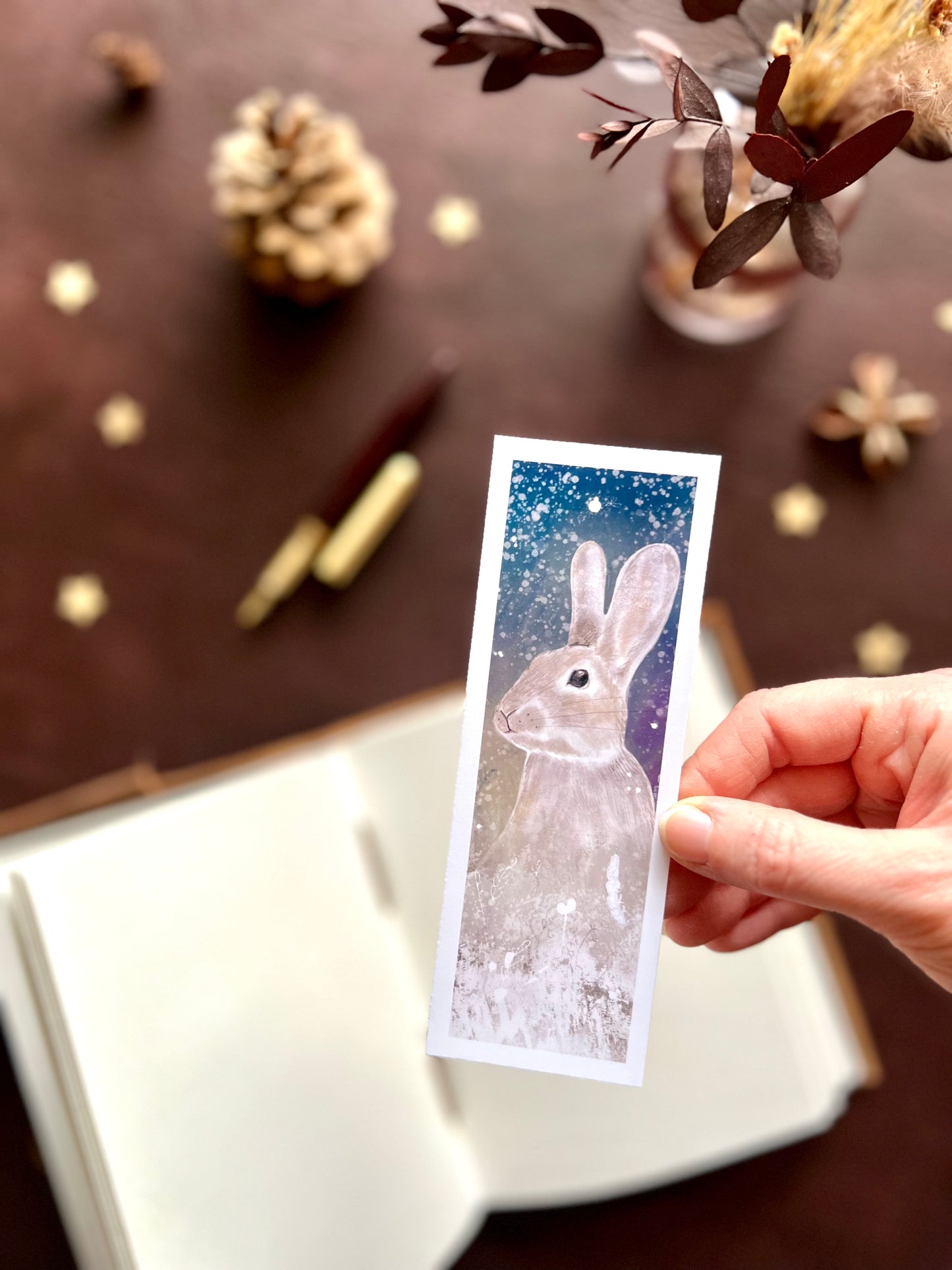 Illustrated bookmark featuring a rabbit beneath a starry night sky in a snowy winter scene. Woodland themed background with sketchbook, pine cones, and kraft paper stars. Part of a 16 pack animal bookmark set for wedding favours.
