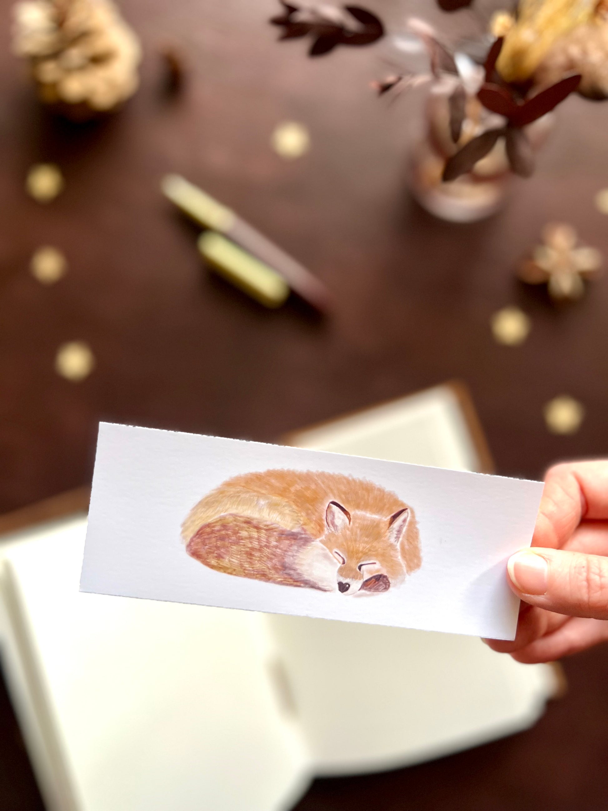 Illustrated bookmark featuring a peaceful fox curled up asleep. Woodland themed background with sketchbook, pine cones, and kraft paper stars. Part of a 16 pack animal bookmark set for wedding favours.
