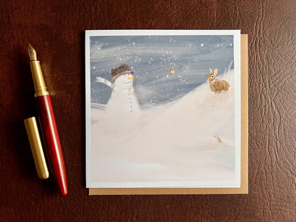 Snowman and Rabbit winter scene greeting card laid flat on table with 110gsm recycled kraft envelope next to wooden fountain pen, UK handmade