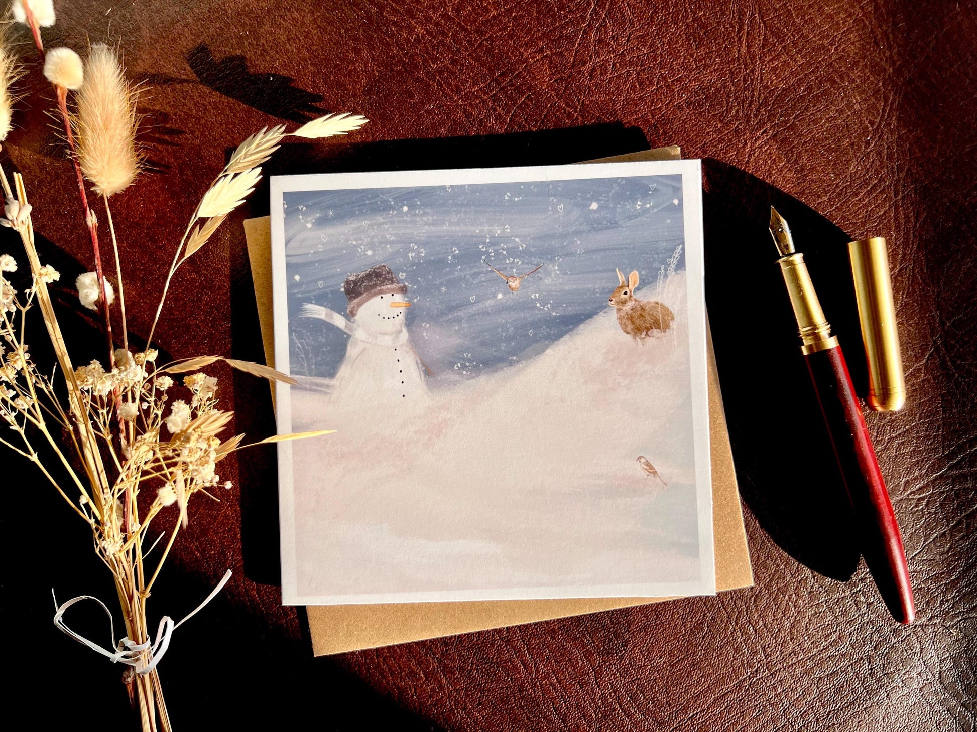 Snowman and Rabbit winter scene greeting card and 110gsm recycled kraft envelope laid flat next to dried flowers and wooden fountain pen, sunlight casting shadows, UK handmade