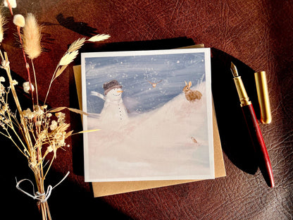 Snowman and Rabbit winter scene greeting card and 110gsm recycled kraft envelope laid flat next to dried flowers and wooden fountain pen, sunlight casting shadows, UK handmade