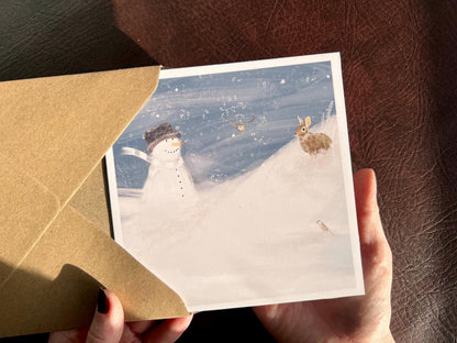 Hands holding a Snowman and Rabbit winter scene greeting card with 110gsm recycled kraft envelope, UK handmade