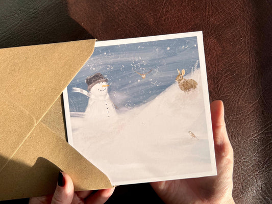 Hands holding a Snowman and Rabbit winter scene greeting card with 110gsm recycled kraft envelope, UK handmade