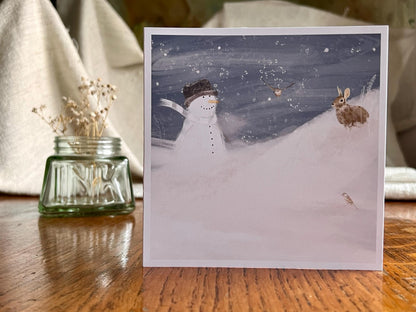 Snowman and Rabbit winter scene greeting card standing on table next to ink pot with dried flowers, UK handmade