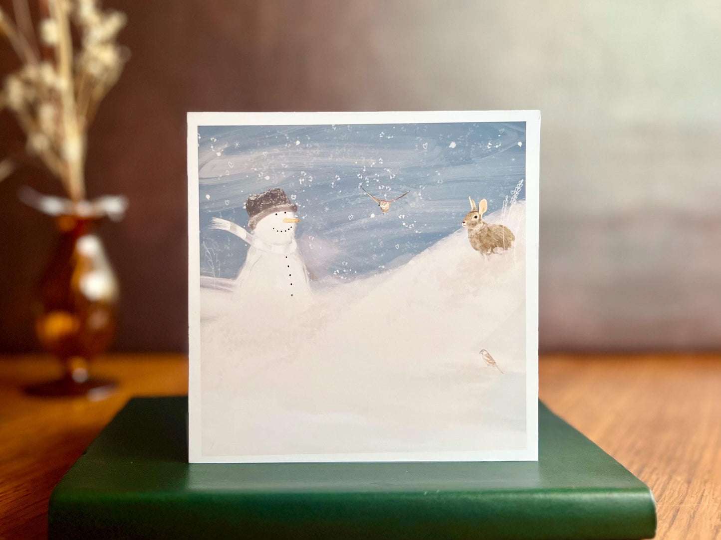Snowman and Rabbit winter scene greeting card standing on green leather book on wooden desk with vase of flowers in background, UK handmade
