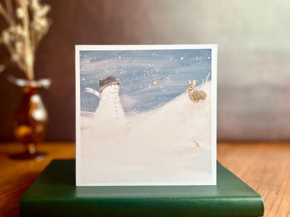 Snowman and Rabbit winter scene greeting card standing on green leather book on wooden desk with vase of flowers in background, UK handmade