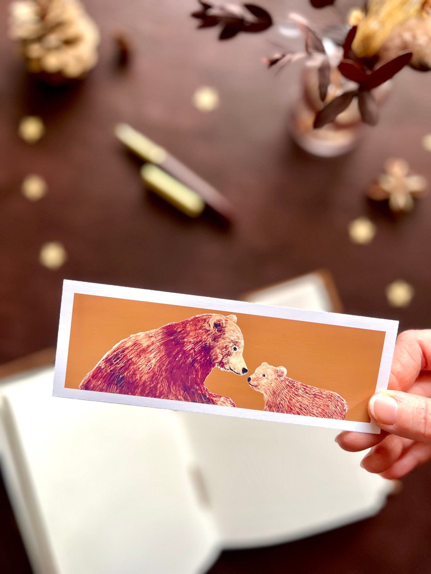 Illustrated bookmark featuring a bear and cub against a warm autumnal background. Woodland themed background with sketchbook, pine cones, and kraft paper stars. Part of a 16 pack animal bookmark set for wedding favours.