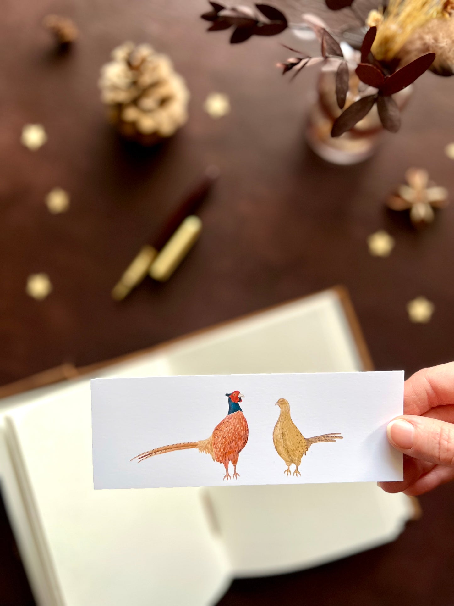 Illustrated bookmark featuring a male and female pheasant standing side by side. Woodland themed background with sketchbook, pine cones, and kraft paper stars. Part of a 16 pack animal bookmark set for wedding favours.