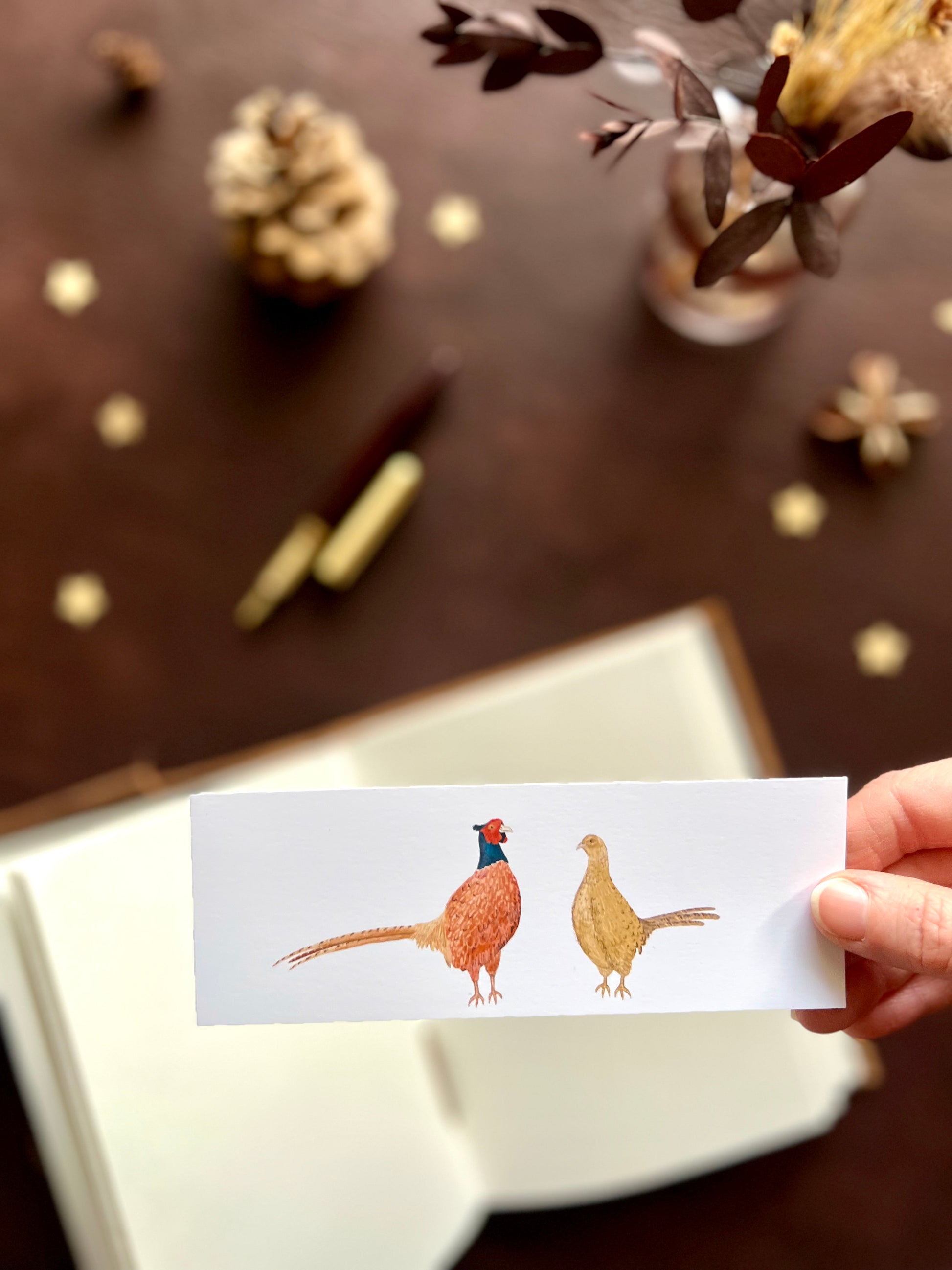 Illustrated bookmark featuring a male and female pheasant standing side by side. Woodland themed background with sketchbook, pine cones, and kraft paper stars. Part of a 16 pack animal bookmark set for wedding favours.