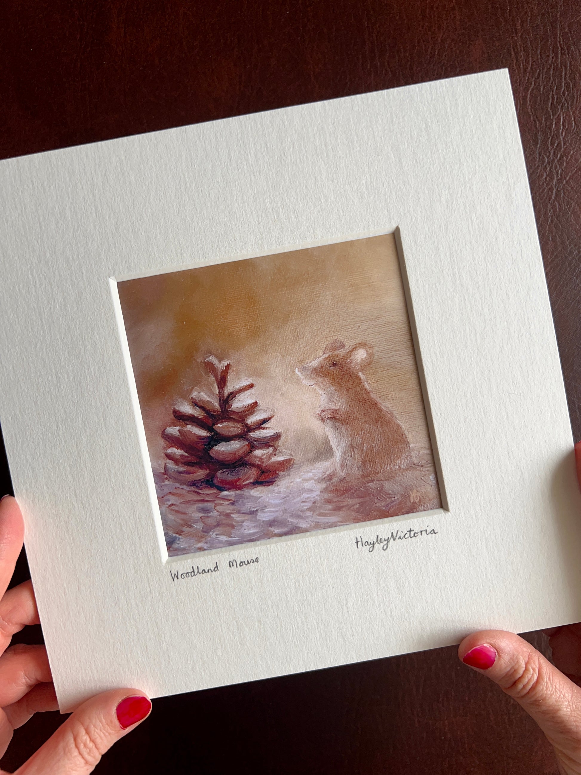Close-up of a mounted Woodland Mouse art print held by the artist, showing the detailed woodland mouse painting, handwritten title and signature, with the angled presentation providing a sense of the artwork's size and texture.