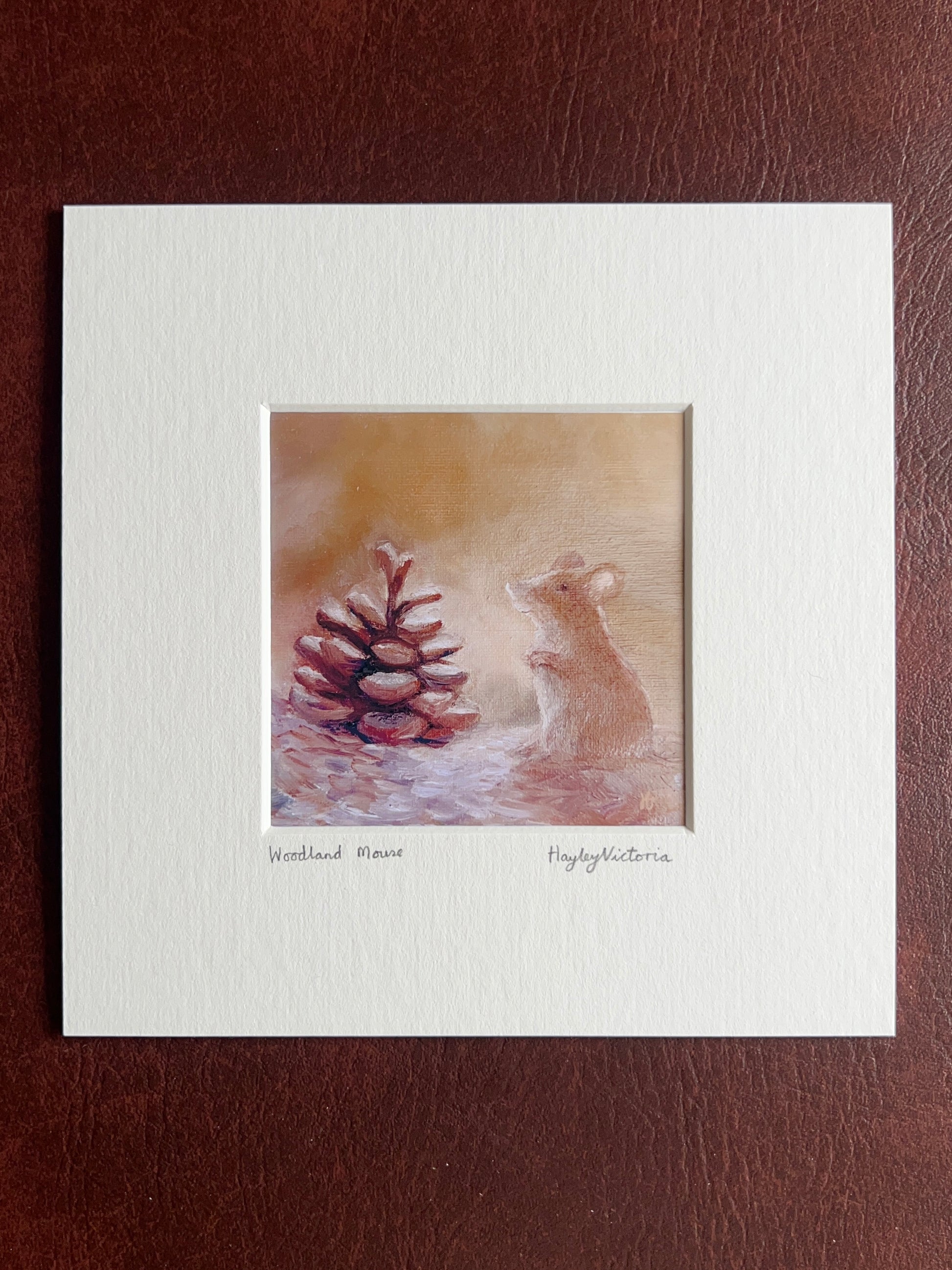 Mounted Woodland Mouse art print displayed on a brown leatherette surface, featuring a warm white textured mount surrounding a whimsical woodland mouse oil painting with handwritten title and artist signature beneath the artwork.
